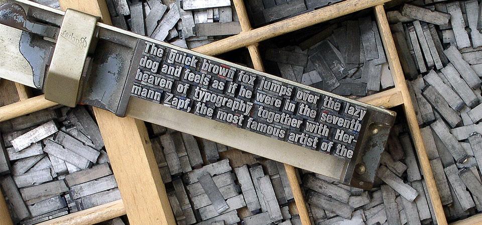 Una caja de piezas tipográficas de metal fundido y material tipográfico en una barra de composición ["El rápido zorro marrón salta sobre el perro perezoso y se siente como si estuviera en el séptimo cielo de la tipografía junto con Hermann Zapf, el artista más famoso de"]