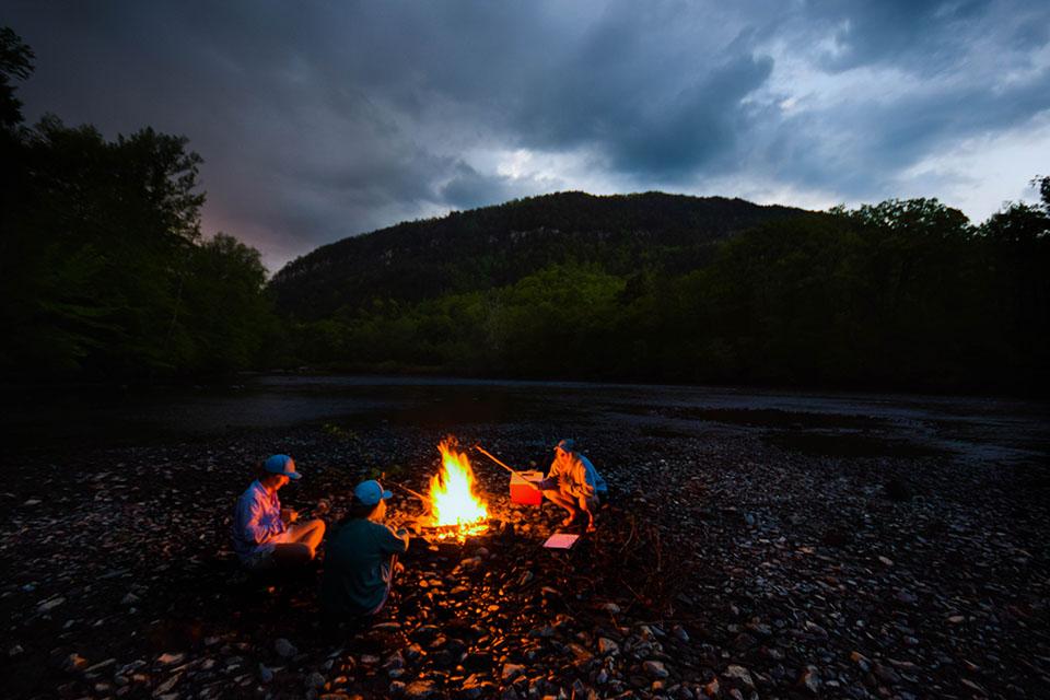 Tres personas rodean una fogata de campamento mientras ponen marshmallows sobre el fuego. La luz anaranjada del fuego iluminando el frente de sus cuerpos, contrasta muchísimo con el azul frío, casi negro, de la noche a su alrededor, bajo el cielo de un atardecer tardío que amenaza lluvia.