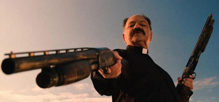 Fotograma de la película Machete, mostrando en contrapicado a un sacerdote mexicano. Es un hombre calvo, con un bigote importante. Con el cielo azul al fondo, el cura tiene un tono de color tirando a ocre mientras encañona a un lado de la imagen con una escopeta y sostiene otra apoyada contra la cadera. Lo que viene siendo un buen personaje de ejemplo de los que son los Tipos Duros.