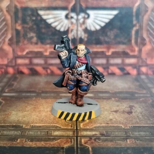 Fotografía de un pistolero clásico de metal de Necromunda. En un escenario de losetas y paredes industriales de metal oxidado, un hombre de piel clara vestido con gabardina oscura, pañuelo rojo al cuello, camiseta azul cielo, pantalones azul oscuro y botas de cuero. Una canana de cartuchos le cruza el pecho, lleva un cinturón y dos pistoleras de cuero, de los que cada tira de cuero de cinturón o ajuste está ocupada por munición. Empuña una pistola bolter en cada mano, con la derecha apuntando al techo y la izquierda abajo, y tiene una pose de avanzar con paso firme.