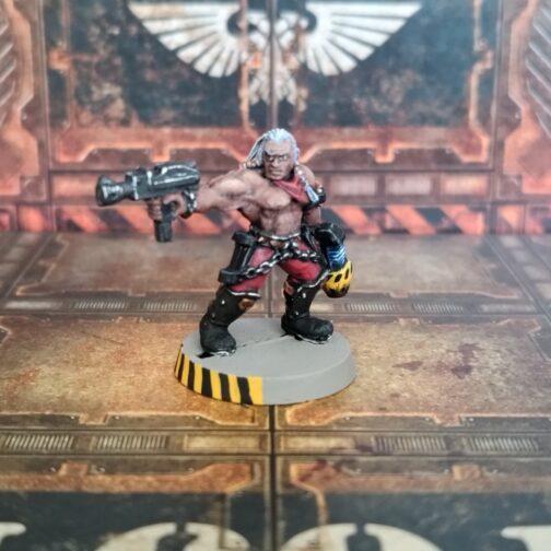 Fotografía de un pistolero clásico de metal de Necromunda. En un escenario de losetas y paredes industriales de metal oxidado, un hombre de piel oscura con el pelo largo blanco con trenzas decoradas por bolitas de metal dorado. Lleva un pañuelo rojo al cuello, desnudo de cintura para arriba, con pantalones rojos, cinturón de acero con tachuelas en bronce del que cuelgan dos pistoleras que se ajustan a los muslos con cadenas de gruesos eslabones. Lleva botas negras con grabas metálicas decoradas con calaveras de bronce a la altura de las rodillas. Está de medio lado, apuntando hacia su derecha con una autopistol mientras sostiene hacia el suelo una pistola de plasma cargada y con el cañón amarillo requemado del uso.