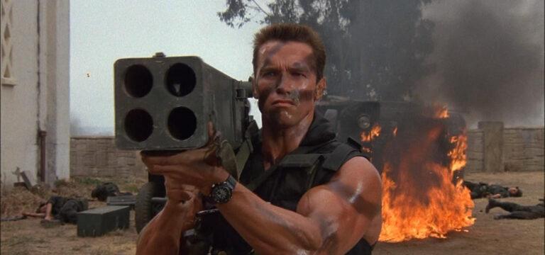 Fotograma de la película Commando. En primer plano está el protagonista, John Matrix, encarnado por Arnold Schwarzenegger, de frente hacia la cámara y empuñando un lanzacohetes cuádruple que apunta hacia delante. Se le ve sólo de mitad del torso para arriba. Viste un chaleco táctico que deja ver los brazos mazadísimos con trazas de camuflaje, como la cara. Detrás de él hay un camión volcado en llamas y montones de cuerpos de soltados.