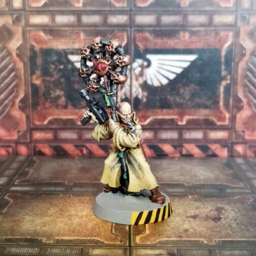 Fotografía de un psíquico clásico de metal de Necromunda. En un escenario de suelos y paredes de aspecto oxidado, un hombre está mirando hacia la derecha de la imagen mientras se inclina hacia la izquierda apoyándose en un gran báculo que coge con la mano izquierda y mantiene levantada una autopistola con la mano izquierda. Es calvo, con una barba canosa hasta casi ser blanca, y la piel muy pálida. Va vestido con un abrigo largo de cuello alto en color caqui, con botas de color cuero claro. El báculo es metálico y está coronado por un círculo vertical de metal gris en cuyo centro hay un ojo rojo rodeado de siete cráneos de color bronce.