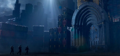 Imagen nocturna, iluminada de forma tenue en tonos azules. Se ve una fachada de un monasterio del Adeptus Mechanicus, con columnas bordeando la puerta. Todos los alrededores y hasta el horizonte está ocupado por montañas de contenedores, formando una geografía artificial de escala planetaria. En medio de la oscuridad, tres formas humanas se dirigen hacia la puerta. Es un fotograma de la serie The Tithes, que podría ser un ejemplo de aventuras en un framework de tier 2 para Wrath and Glory Warhammer 40000 Roleplay.