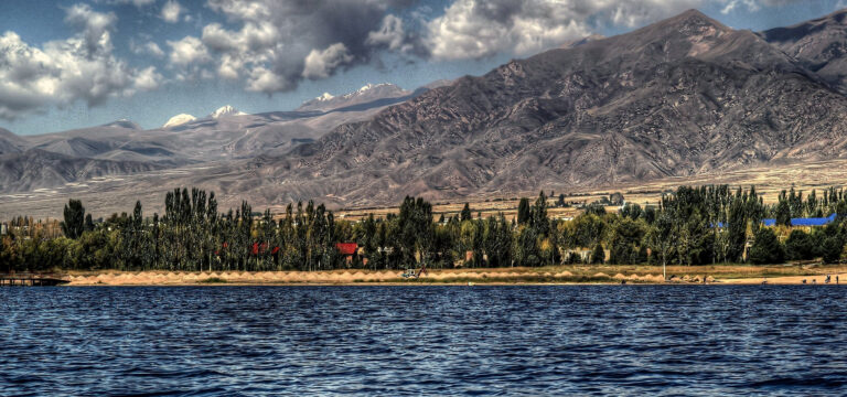Fotografía del litoral norte del lago Issyk en Kirguistán. Las aguas son intensamente azules, mientras que la tierra es muy clara y está jalonada por parches de árboles altos y estrechos. El horizonte entero está compuesto por montañas irregulares de un color gris ceniza. Es una fotografía de Thomas Depenbush, compartida bajo licencia CC2.0. Un lugar propio de curiosidades históricas.