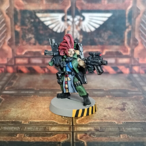 Fotografía frontal en escorzo de un cazarrecompensas clásico de metal de Necromunda. En un escenario de losetas y paredes industriales de metal oxidado, un hombre de pelo largo de un rojo intenso peinado hacia arriba con un estilo entre cardado y punk. Lleva un uniforme militar de color verde, con un tabardo que le cae entre las piernas en color azul militar cubierto de sellos de pureza. Bajo la chaqueta lleva una camiseta azul brillante y está calzado con botas militares negras. Empuña en su mano izquierda una pistola bolter negra con una mira de lentes rojas, con la que apunta hacia la derecha de la imagen, mientras en la izquierda sostiene un rifle bolter de cuerpo negro apoyado sobre la cadera. Va equipado con una máscara respiradora colgada del cuello y una mochila a la espalda de la que sobresalen una escopeta y la empuñadura de una espada.