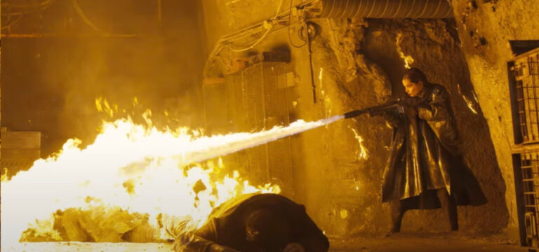 En un entorno de paredes con aspecto de hormigón industrial, con armarios, cables y tuberías de metal oxidado, una chica con un abrigo ignífigo negro dispara un lanzallamas sobre dos cuerpos tendidos en el suelo. Toda la escena está iluminada en el intenso color amarillo anaranjado del fuego. Es de una película, pero también es algo típico de Necromunda para las armas con el rasgo Blaze.