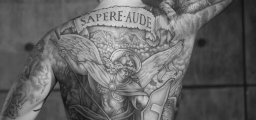 Fotografía en blanco y negro de la espalda completamente tatuada de un hombre. El tatuaje es una hueste angelical, con todo el centro de la espalda ocupada por un único ángel con escudo y lanza, cargando hacia abajo. Un pergamino en el tatuaje, bajo el cuello, tiene el texto "SAPERE AUDE". El autor del tatuaje es Anton Ivkin y la fotografía es de Alexander Kuzovlev, siendo libre de derechos. Nos sirve para ilustrar la forma de redefinir la magia en La Marca del Este y juegos OSR por medio de tatuajes volubles.
