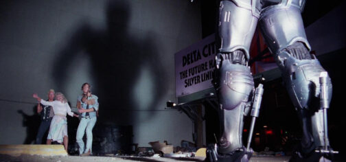 Fotograma de la película Robocop de 1987. En primer plano se ven las piernas blindadas de Robocop, desde atrás que es también desde donde le iluminan los faros del coche proyectando una sombra enorme sobre la pared del edificio del fondo. Iluminados justo frente a la pared y enfrentados a Robocop con una navaja, se encuentran dos delincuentes que han atrapado a una mujer. Un ejemplo de lo que representa el mundo de Tipos Duros.