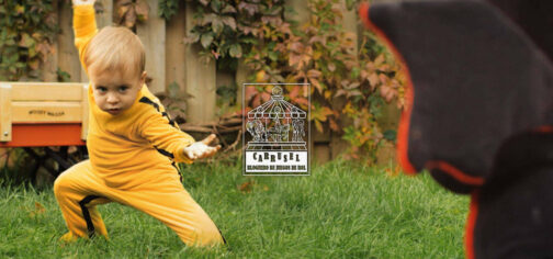 En un jardín de casa con césped alto, un bebé rubio vestido con un mono como el de La Novia de Kill Bill se pone en posición de combate frente a un peluche. No es un ejemplo de baja letalidad en campañas de rol, pero es gracioso. En el centro sobre la fotografía está el logo de la iniciativa Carrusel: un rectángulo vertical con un tiovivo de feria coronado por un dado de 20 caras, cuyas monturas son una imagen de Cthulhu, un caballero armado a caballo y una armadura de combate tecnológica. Bajo el tiovivo aparecen las letras grandes CARRUSEL ondulando arriba y abajo, y bajo esas, ocupando el mismo ancho, BLOGUERO DE JUEGOS DE ROL.