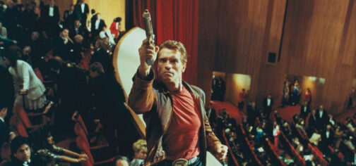 Jack Slater, el protagonista de El Último Gran Héroe, en la escena de la ópera apuntando con su pistola Desert Eagle hacia los palcos superiores. Es el actor Arnold Schwarzenegger, vestido con chaqueta de piel desgastada, camiseta roja y pantalones vaqueros con una hebilla grande como un piano.