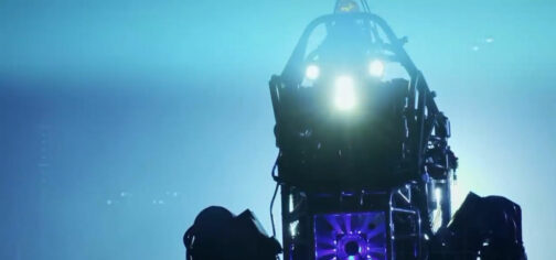 En un fondo saturado de diferentes fuentes de luz azul se recorta la estructura de tubos metálicos, ventilación iluminada en morado y los tres focos de luz de un robot de DARPA, dándole un aspecto un poco de película de terror.