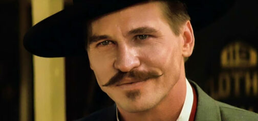 Primer plano de Val Kilmer como Doc Hollyday en Tombstone, con camisa blanca, chaleco rojo, camisa verde y sombrero negro. Es un personaje que cuando participó en el famoso duelo en OK Corral ya estaba muy afectado por la tuberculosis y sabía que en cualquier momento podía empezar el último viaje, como la desventaja de Savage Worlds para Tipos Duros.