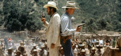 Fotograma de la película Traición sin límites (Extreme Prejudice), de 1987. Hay dos hombres espalda contra espalda en el centro, mientras alrededor se reúnen montones de secuaces de uno de ellos en lo que parece el típico pueblo tercermundista que el EEUU piensan que es México. El de la izquierda es Cash Bailey, a la derecha y vestido con traje blanco sucio, sombrero blanco y un revolver de cañón corto. El de la derecha es Jack Benteen, con sombrero blanco con cinta negra, camisa azul clara y pantalón vaquero oscuro, empuñando un Colt 45. Parecen a punto de batirse en duelo. Son dos tipos de fuertes convicciones, aunque opuestas entre sí.