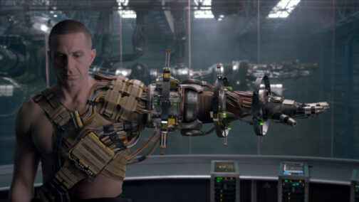 Forograma de la película Pacific Rim en la que se ve a un hombre, desnudo de cintura para arriba y con el pelo rapado, con el brazo izquierdo extendido por completo. Lleva un arnés lleno de cables, sensores y baterías que se extiende a lo largo de varios soportes y articulaciones alrededor del brazo.