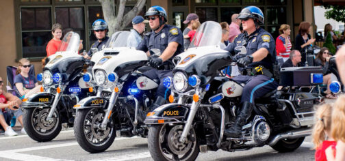 Tres policías montados en motocicletas de gran cilindrada, con sus uniformes azules oscuros con una raya azul más clara en el pantalón y el casco. Parecen estar en medio de un desfile, porque hay gente observándolos desde ambos lados de una calle.