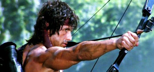 John Rambo, con el pelo desgreñado sostenido por su cinta roja, sin camiseta y cubierto de suciedad, apunta con su arco de poleas cargado con una de sus famosas flechas explosivas en medio de la selva. De esas flechas explositvas tenía que haber en Tipos Duros y Savage Worlds.
