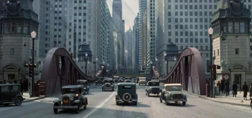 Vista de los años 30 del puente de la calle Clark en Chicago. Es una estructura metálica diseñada para dividirse y elevarse, permitiendo el tráfico fluvial, que en estos momentos se encuentra cerrado y por el que circulan un montón de coches de modelos de los años 30 y en cuya prolongación se pueden ver grandes bloques de edificios de hormigón gris.