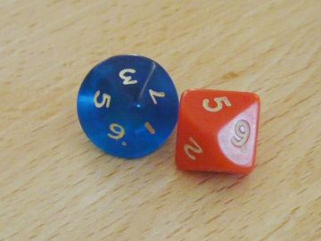 Los 2d10 originales con los que empezó todo. El génesis del frikismo. Es una fotografía de mala calidad de un primer plano de ambos dados sobre un tablero de madera clara. El de la izquierda es azul oscuro, traslúcido, con letras blancas, mostrándose casi "de punta" hacia la cámara de forma que se ve prácticamente circular mientras muestra sólo las caras de los resultados impares con un resultado de 3. El de la derecha es naranja butano con letras blancas y está en una posición más de lado, un poco en la forma que se suele representar icónicamente el d10, mostrando el resultado de 5 y otras dos caras legibles con el 9 y el 2.