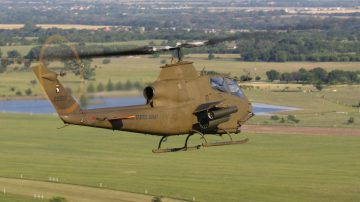 Vista desde el ángulo atrás a la derecha de un Bell TAH-1p Huey Cobra. Es un helicóptero de ataque del US Army, de color verde-marrón militar, monomotor equipado con dos lanzacohetes y un cañón rotatorio de 30mm en el morro, volando sobre campiña y bosques. Es uno de los tipos helicópteros militares para Savage Worlds contemplados en Tipos Duros. La fotografía del 1976 Bell TAH-1P Huey Cobra 76-22599 - N599HF, Vietnam War Flight Museum es de Christopher Ebdon con licencia CC BY-NC-SA 2.0.