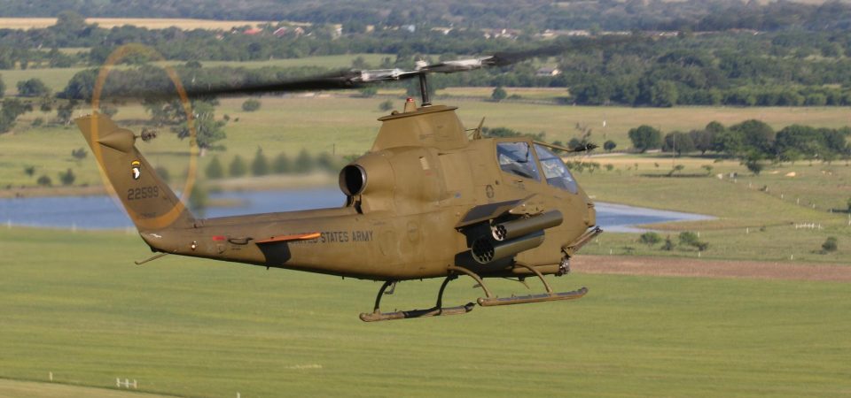 Vista desde el ángulo atrás a la derecha de un Bell TAH-1p Huey Cobra. Es un helicóptero de ataque del US Army, de color verde-marrón militar, monomotor equipado con dos lanzacohetes y un cañón rotatorio de 30mm en el morro, volando sobre campiña y bosques. Es uno de los tipos helicópteros militares para Savage Worlds contemplados en Tipos Duros. La fotografía del 1976 Bell TAH-1P Huey Cobra 76-22599 - N599HF, Vietnam War Flight Museum es de Christopher Ebdon con licencia CC BY-NC-SA 2.0.