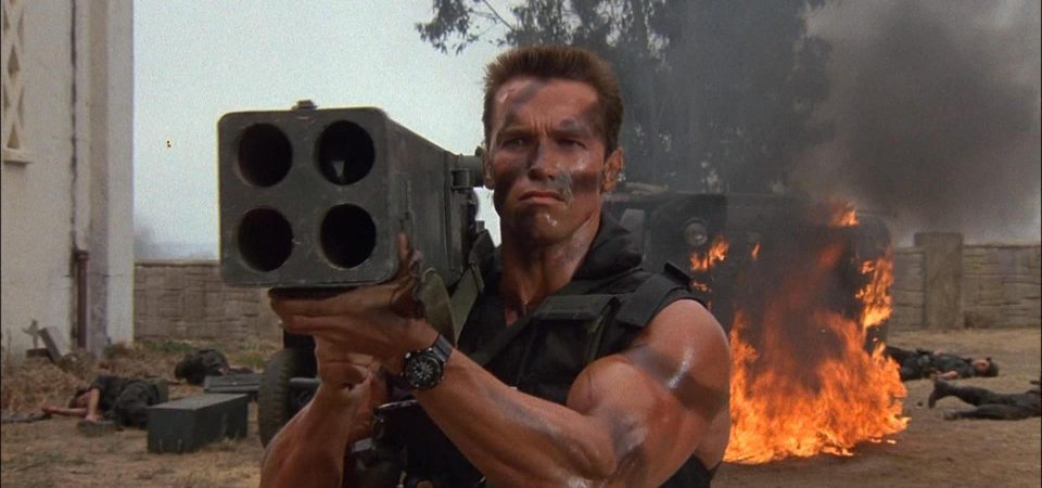 Fotograma de la película Commando. En primer plano está el protagonista, John Matrix, encarnado por Arnold Schwarzenegger, de frente hacia la cámara y empuñando un lanzacohetes cuádruple que apunta hacia delante. Se le ve sólo de mitad del torso para arriba. Viste un chaleco táctico que deja ver los brazos mazadísimos con trazas de camuflaje, como la cara. Detrás de él hay un camión volcado en llamas y montones de cuerpos de soltados.