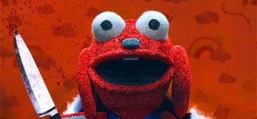 Primer plano de la cara de Benny, el arrollador protagonista absoluto de la película slasher Benny Loves You (Benny te ama). Es un peluche de color rojo intenso, con nariz achatada negra, enormes ojos saltones que sobresalen por encima de la frente, la boca abierta en lo que resulta una sonrisa mona y una tira de trapo haciendo las veces de sus dos únicos dientes, las paletas de arriba. A los lados de la cabeza le cuelgan dos largas orejas como las de un perro orejón. Es un peluche de aspecto feo pero simpático a pesar de que se ve que sostiene un cuchillo ensangrentado.