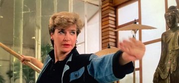 Fotograma de una película de Cynthia Rothrock. Es una mujer rubia, con el pelo muy corto, de ojos azules, lleva una chaqueta de corte ochentero en color azul vaquero y negro. Empuña una vara en la mano derecha y adelanta la izquierda en una pose de espera de artes marciales. La imagen trata de representar Elizabeth McCray, un personaje del juego de rol Tipos Duros, en el universo Wildspring, que la entrada describe como un personaje pregenerado de Savage Worlds.