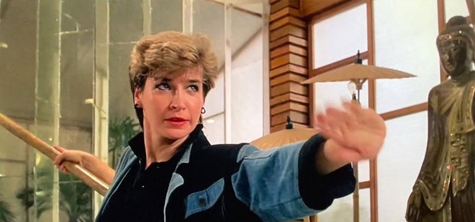 Fotograma de una película de Cynthia Rothrock. Es una mujer rubia, con el pelo muy corto, de ojos azules, lleva una chaqueta de corte ochentero en color azul vaquero y negro. Empuña una vara en la mano derecha y adelanta la izquierda en una pose de espera de artes marciales. La imagen trata de representar Elizabeth McCray, un personaje del juego de rol Tipos Duros, en el universo Wildspring, que la entrada describe como un personaje pregenerado de Savage Worlds.