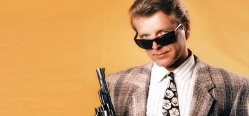 Fotografía de David Rasche, mirando a cámara, con las gafas de sol cayendo sobre la nariz sin cubrir los ojos, con una americana a cuadros escoceses, una camisa a rayas blancas verticales blancas y gris perla, una corbata azul con flores blancas y empuñando un revólver, que apunta hacia arriba. Es el actor que se quiso para hacer de Norman Jackson en el casting de Wildspring, la serie que adapta Tipos Duros. La imagen se utiliza para ilustrar el aspecto de Norman Jackson como personaje para Savage Worlds.