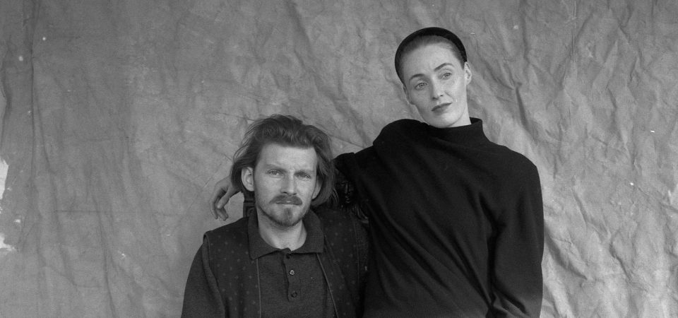 Fotografía en blanco y negro de Brendan Perry y Lisa Lisa Gerrard, los componentes de Dead Can Dance. Él está sentado en una silla (fuera de plano) y lleva un polo con un chaleco estampado por encima, con barba poco poblada y bigote más marcado, así como el pelo negro, mientras mira a cámara. Ella parece estar sentada sobre él, pasándole un brazo por encima del hombro, mientras mira hacia el lado; lleva el pelo recogido bajo un gorro ceñido y viste un jersey.