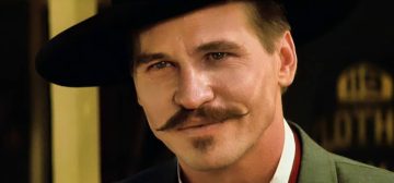 Primer plano de Val Kilmer como Doc Hollyday en Tombstone, con camisa blanca, chaleco rojo, camisa verde y sombrero negro. Es un personaje que cuando participó en el famoso duelo en OK Corral ya estaba muy afectado por la tuberculosis y sabía que en cualquier momento podía empezar el último viaje, como la desventaja de Savage Worlds para Tipos Duros.