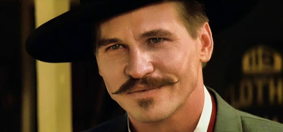 Primer plano de Val Kilmer como Doc Hollyday en Tombstone, con camisa blanca, chaleco rojo, camisa verde y sombrero negro. Es un personaje que cuando participó en el famoso duelo en OK Corral ya estaba muy afectado por la tuberculosis y sabía que en cualquier momento podía empezar el último viaje, como la desventaja de Savage Worlds para Tipos Duros.