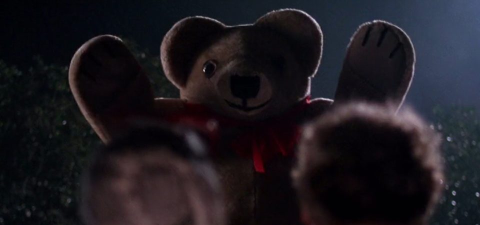Un oso de peluche de un gran tamaño se yergue en una noche oscura, mirando hacia el espectador. Hay poca luz, pero muy contrastada, por lo que las facciones del peluche y sus contornos apenas se ven perfiladas. En primer plano, de espaldas, hay dos cabezas desenfocadas de las personas que están viendo al peluche. La imagen en sí presenta una imagen tan onírica como terrorífica, al mostrar al peluche de manera anormalmente grande y amenazador. Es un fotograma de la película Dolls, de Stuart Gordon del año 87, pero podría ser de la aventura de juego de rol La noche sombría de los muñecos.