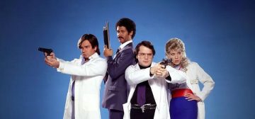 Imagen promocional de la serie Garth Marenghi's Darkplace. Los cuatro protagonistas de esta serie B se encuentran sobre un fondo azul que es más claro a su alrededor y más oscuro hacia los extremos. De derecha a izquierda, encontramos a: el doctor Lucien Sánchez, con su bata blanca, girado hacia la izquierda mientras apunta con una pistola; Dean Lerner, con traje azul oscuro y camisa blanca, mirando directamente hacia el espectador y sosteniendo una escopeta con el cañón hacia el techo; el doctor Rick Dagless M.D., con la bata blanca sobre pantalón y camisa negra, cinturón con hebilla de calavera de cuernilargo en plata, corbata morada y apuntando hacia la izquierda con un revólver; la doctora Madeleine Wool, con el cabello rubio cardado en un estilo muy ochentero, y la bata blanca sobre una camisa a rayas azules, un ceñidor de cintura rojo intenso y falda de tubo azul (es la única que no empuña un arma).