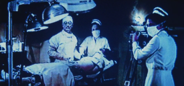 Fotograma de baja calidad y con mucho grano, en monocromo de tonos azules, que muestra un quirófano mugriento de mediados del S. XX en el que una enfermera graba con una cámara antigua a un tipo que está atado a la camilla mientras un médico con expresión perturbadora, y sin mascarilla, y una segunda enfermera están a su lado sosteniendo material quirúrgico. Los tres personajes que no están atados a la camilla miran a cámara con expresión sorprendida. El tipo de situación que nadie quisiera encontrar durante una evaluación de proyecto.