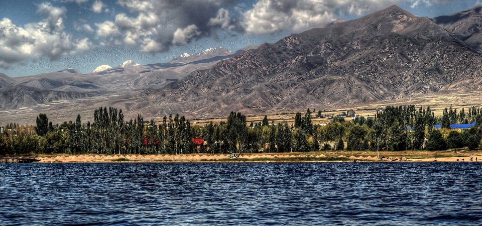 Fotografía del litoral norte del lago Issyk en Kirguistán. Las aguas son intensamente azules, mientras que la tierra es muy clara y está jalonada por parches de árboles altos y estrechos. El horizonte entero está compuesto por montañas irregulares de un color gris ceniza. Es una fotografía de Thomas Depenbush, compartida bajo licencia CC2.0. Un lugar propio de curiosidades históricas.