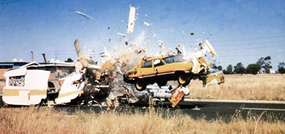 Un coche Ford Falcon de 1974 de color amarillo con una franja roja y otra blanca que lo cruzan de lado a lado, y la parte de abajo en azul, atraviesa un remolque caravana, haciéndolo pedazos y lanzando fragmentos de ésta en todas direcciones. Esto ocurre en medio de una carretera recta rodeada de hierba seca. Es un fotograma de la película Mad Max, en la que se ven muchos ejemplos de eso de estrellar vehículos en Savage Worlds.