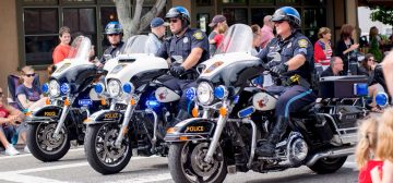 Tres policías montados en motocicletas de gran cilindrada, con sus uniformes azules oscuros con una raya azul más clara en el pantalón y el casco. Parecen estar en medio de un desfile, porque hay gente observándolos desde ambos lados de una calle.