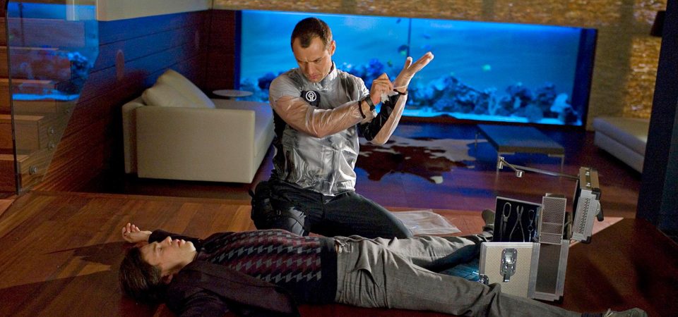 Fotograma de la película Repo Men. En un escenario que parece un apartamento de lujo, hay un tipo inconsciente sobre el suelo de parquet y Jude Law está arrodillado a su lado mientras lo observa con detenimiento y se ajusta unos guantes quirúrgicos.