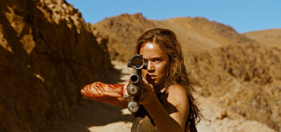 Fotograma de la película Revenge. En un paraje desolado, de tierra ocre, sin vegetación alguna, una chica con mirada de absoluta determinación, con pelo largo y camiseta sin mangas, el brazo izquierdo y la frente ensangrentadas, encañona hacia el espectador con una escopeta con mira telescópica. Ilustra un fotograma de la película perdida Task Force Final Girl, del trasfondo del juego de rol Tipos Duros.