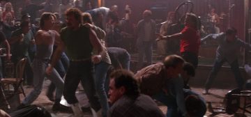 Una multitudinaria pelea de bar de la película Road House de 1989. Docenas de tipos en un local iluminado apenas a media luz, con el ambiente muy cargado, se zurran entre ellos sin orden ni concierto en una melee descontrolada por completo. Es un caos de puñetazos, botellazos y sillazos. Un clásico candidato a la resolución rápida de escenas de acción con el reglamento de Savage Worlds y Tipos Duros.