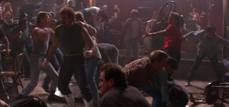 Una multitudinaria pelea de bar de la película Road House de 1989. Docenas de tipos en un local iluminado apenas a media luz, con el ambiente muy cargado, se zurran entre ellos sin orden ni concierto en una melee descontrolada por completo. Es un caos de puñetazos, botellazos y sillazos. Un clásico candidato a la resolución rápida de escenas de acción con el reglamento de Savage Worlds y Tipos Duros.