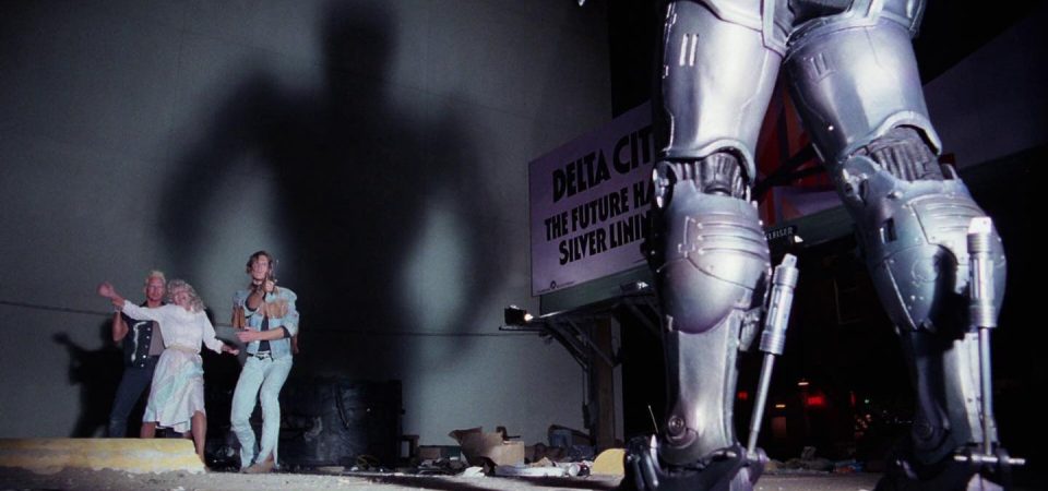 Fotograma de la película Robocop de 1987. En primer plano se ven las piernas blindadas de Robocop, desde atrás que es también desde donde le iluminan los faros del coche proyectando una sombra enorme sobre la pared del edificio del fondo. Iluminados justo frente a la pared y enfrentados a Robocop con una navaja, se encuentran dos delincuentes que han atrapado a una mujer. Un ejemplo de lo que representa el mundo de Tipos Duros.