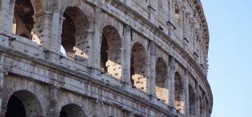 Fotografía de la fachada del Coliseo de Roma, en contrapicado, de cerca y con el detalle de sus últimos dos pisos de manera que se ven muy bien sus arcos y decoración, mientras la perspectiva permite que se recorte la circunferencia contra el cielo azul completamente claro.
