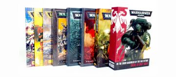 Fotografía de los manuales de seis ediciones de Warhammer 40000, superpuestos de más antigua a más reciente. Los tres más antiguos son los tres manuales de Segunda Edición, y va pasando por tercera, cuarta, quinta y sexta hasta que la más visible es la caja contenedora de los manuales de séptima.