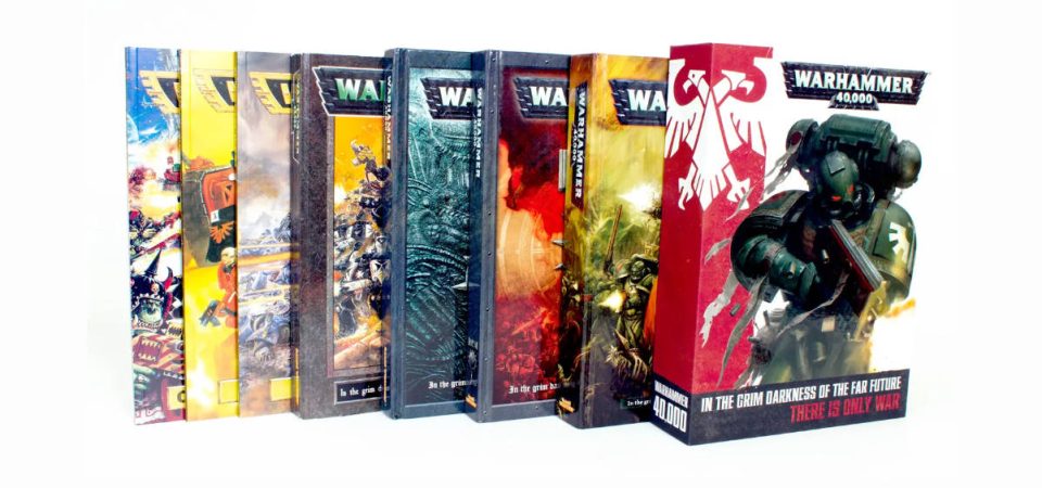 Fotografía de los manuales de seis ediciones de Warhammer 40000, superpuestos de más antigua a más reciente. Los tres más antiguos son los tres manuales de Segunda Edición, y va pasando por tercera, cuarta, quinta y sexta hasta que la más visible es la caja contenedora de los manuales de séptima.