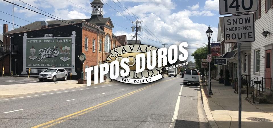 Una calle cualquiera de una pequeña ciudad de estados unidos, con edificios de ladrillo rojo, madera blanca y varios coches circulando o aparcando a los lados de la amplia calzada. Podría ser una calle cualquiera de la serie Wildspring, en la que los personajes de Savage Worlds viven las aventuras de Tipos Duros. Fotografía tomada por Famartin con licencia CC BY-SA 4.0. Sobre la imagen está impreso el logo de Tipos Duros, el juego de rol: el texto Tipos Duros en letras blancas grandes y cuadradas, con una inclinación de unos 15º hacia la derecha y debajo, letra más pequeña pero con las mismas características Edición Aventura, todo girado unos 15º a contrarreloj y superpuesto al logo de Savage Worlds Fans Product.