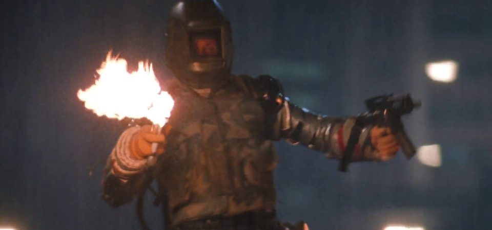 Un hombre vestido con un traje ignifugo plateado cubierto de placas de blindaje caseras y una máscara facial de armadura hecha a medida, empuñando un fusil de asalto en la mano izquierda y un lanzallamas casero en la mano derecha. Lleva puesto un chaleco táctico en colores de camuflaje a pesar de que se encuentra en medio de la ciudad. Es un perfecto ejemplo de tipo que se cree un tanque en Tipos Duros para Savage Worlds.