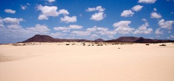 Fotografía de un desierto español. En el centro de la imagen se ve una cordillera montañosa de baja altura y color roca oscura. Debajo la arena de un tono muy claro y sobre las montañas un cielo claro con una multitud de pequeñas nubes blancas. La imagen es CC de Dirk Vorderstrasse.